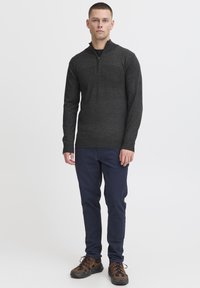 Tamno siva zip-up dukserica s visokim ovratnikom, prilagođenog kroja, u kombinaciji s tamnoplavim hlačama i smešanim patikama s uzorkom. Jednobojna tkanina, pletena tekstura.
