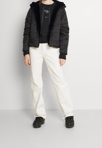 Svart vadderad jacka med huva av fuskpäls, dragkedja framtill, bärs över en svart sweatshirt och vita vida byxor, kombinerat med svarta sneakers.