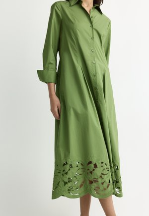 Green long-sleeve button-up dress with collar, pleats, and floral cut-out designs along the hem, worn by a person seen from shoulders to calves.