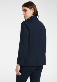 Mujer con cabello castaño que lleva pendientes plateados, de perfil, vestida con un blazer azul marino oscuro y pantalones a juego, sobre un fondo liso.