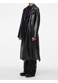 Trench-coat en cuir noir à finition brillante, à double boutonnage, avec de larges revers et des manches longues, porté sur un pantalon noir ample et texturé.