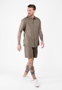 Homme avec des tatouages sur les bras et les jambes, portant une chemise à manches courtes marron à boutons, un short assorti et des baskets blanches, marchant et regardant à gauche.