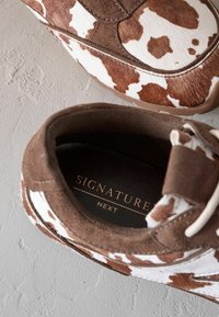 Ruskeat ja valkoiset kuvioidut lenkkarit, joissa on mokka- ja turkispintaa, varustettuna vaaleanruskeilla nauhoilla sekä mustalla sisäpuolella ja kultaisella brändäyksellä.