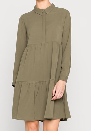 Femme portant une robe longue à volants vert olive à manches longues, avec un col pointu et des boutons dissimulés sur le devant, debout devant un fond uni.