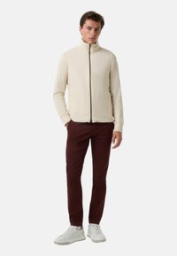 Helle beige Zip-Jacke mit hohem Kragen, langen Ärmeln und Seitentaschen, kombiniert mit burgunderroten Hosen und weißen Sneakers.