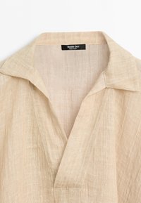 Béžová lněná košile s otevřeným límcem a výstřihem do V, s černou štítkem Massimo Dutti uvnitř límce.