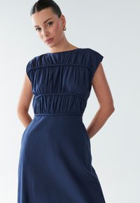 Tamsiai mėlyna suknelė su trumpomis rankovėmis, pasižyminti suvertu liemeniu ir lygiu sijonu. Audinys yra minkštas ir šiek tiek blizgus.