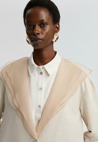 Béžová oversize bunda s textúrovaným, vrstveným golierom. Pod ňou biela košeľa s gombíkmi. Model má na sebe výrazné zlaté kruhové náušnice.
