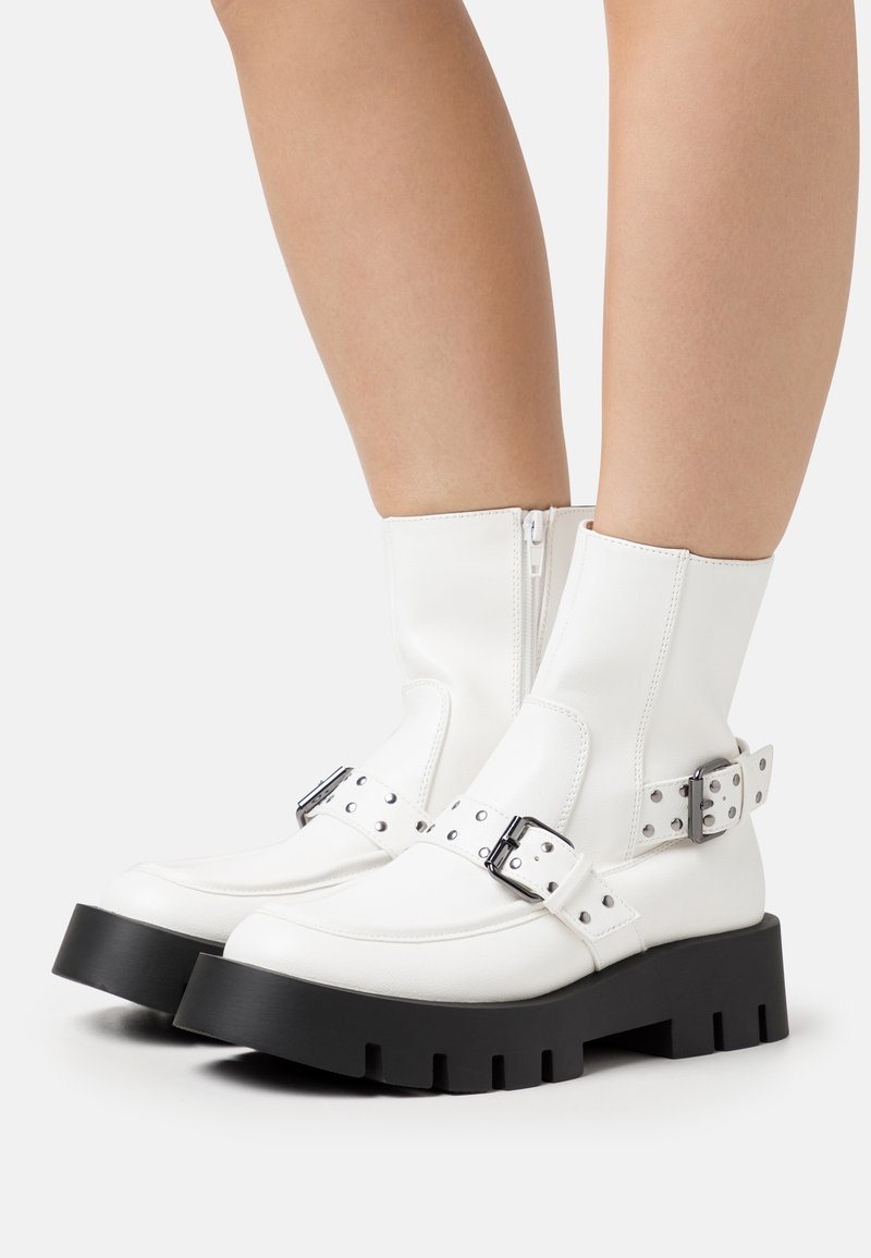 Bottes blanches mi-mollet avec semelles noires épaisses et boucles argentées, portées sur des jambes nues sur un fond uni.