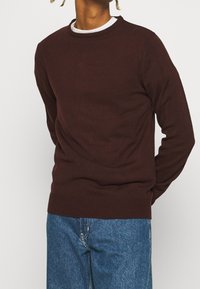 Bordeauxfärgad stickad tröja med rund halsringning och ribbade muddar, bärs över en vit skjorta, ihop med blå denimjeans. Enkel design, mjuk textur.