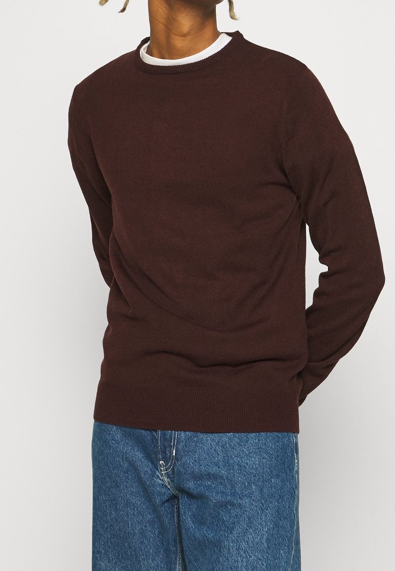 Bordeauxfärgad stickad tröja med rund halsringning och ribbade muddar, bärs över en vit skjorta, ihop med blå denimjeans. Enkel design, mjuk textur.