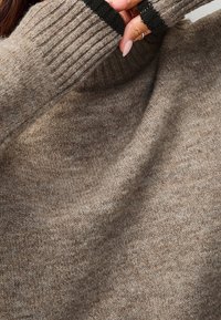 Pull en tricot de couleur brun clair avec un col roulé côtelé et des accents noirs sur les poignets. Texture douce et coupe décontractée.