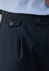 Pantalones ajustados de un azul oscuro, hechos de un tejido texturizado, que presentan dos botones marrones, pliegues y un diseño de frente plano.
