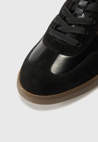 Sneaker nera con tomaia in suede, dettagli in pelle lucida e suola in gomma marrone texturizzata. Presenta lacci neri e un design della punta arrotondata.