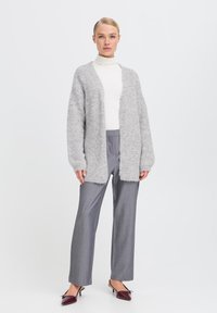 Cardigan grigio chiaro, morbido e fluffy, con apertura frontale, maniche lunghe e senza chiusure, indossato sopra un dolcevita bianco e pantaloni grigi.