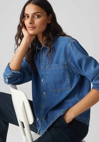 Camicia di jeans in blu, con chiusura a bottoni, taschino singolo sul petto e maniche arrotolate. Il tessuto è liscio con un taglio classico.