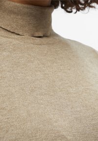 Pull à col roulé tan en tissu tricoté doux, avec une coupe ajustée et une texture à côtes fines. Aucun motif ou accent visible.