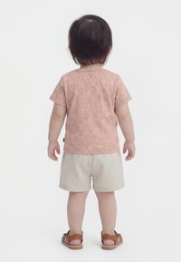 Taapero seisoo selinpäin, päällään vaaleanpunainen kuviollinen paita, beiget shortsit ja ruskeat sandaalit valkoista taustaa vasten.