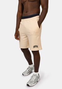 Beige katoenen shorts met elastische tailleband en zijzakken. Voorzien van zwart "CRYSTAL" textlogo op het linkerpijp. Gepaard met zilveren sneakers.