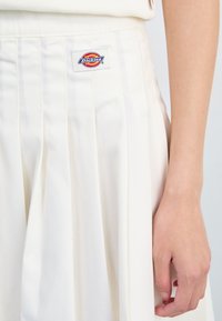 Hvit plisserte skjørt laget av stoff med en glatt tekstur. Har et sydd logo-patch på midjebåndet.