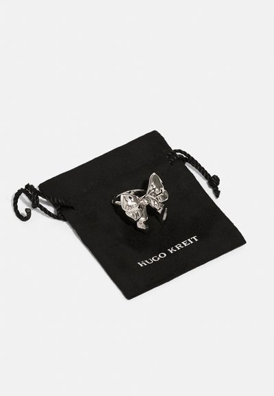 Anello in argento a forma di farfalla con finitura lucida, posizionato su una borsa in tessuto nero con cordoncino e etichetta stampata "HUGO KREIT".