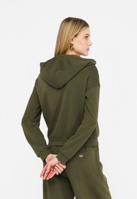 Felpa verde oliva con tasca frontale, cappuccio con cordino e polsini a costine. Tessuto liscio, design morbido, abbinata a pantaloni coordinati.