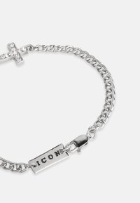 Stříbrný řetízkový náramek s křížem posetým kamínky a štítkem s vyrytým nápisem "ICON." Má pevné zapínání a hladký povrch.