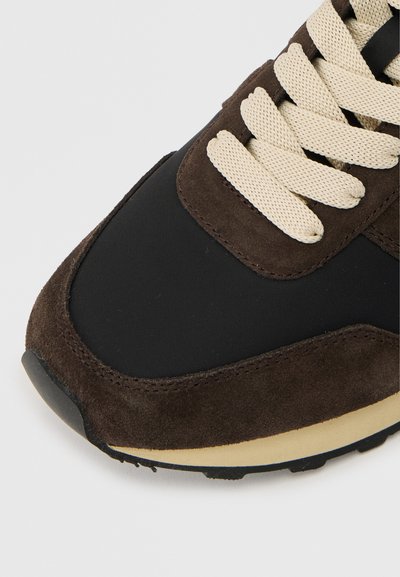 Κοντινό πλάνο στο μπροστινό μέρος ενός αθλητικού παπουτσιού από σκούρο καφέ σουέντ και μαύρο ύφασμα με παχιές κρεμ κορδόνια και μπεζ ενδιάμεση σόλα.