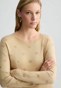 Maglione beige a maglia con scollatura tonda, maniche lunghe e un motivo a logo in rilievo di un colore tan contrastante. Orecchini a forma di cuore dorati.