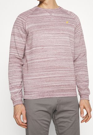 Mand iført en langærmet sweater med rund hals og vandrette lyserøde og hvide striber samt grå bukser.