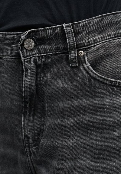 Κοντινό πλάνο της μέσης μαύρου τζιν με μεταλλικό κουμπί και θηλιά ζώνης, φορεμένο πάνω από μαύρο πουκάμισο.