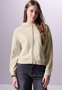Sudadera con cremallera de color verde claro, fabricada en tejido de punto tipo waffle con textura, hombros caídos y un bolsillo frontal. Combinada con jeans de color gris oscuro, diseño sencillo.