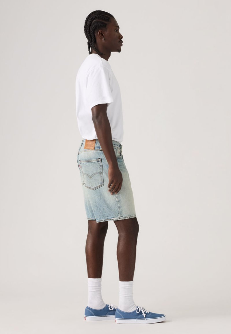Gonna di jeans in lavaggio chiaro con orlo grezzo, abbinata a una T-shirt bianca e sneaker blu, con calzini bianchi e una targhetta in pelle sul retro.