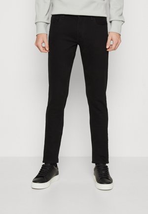 Homme portant un jean noir slim, des baskets basses noires avec des semelles blanches, et une chemise à manches longues gris clair, debout devant un fond blanc uni.