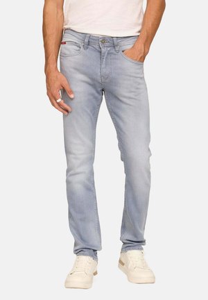 Homme portant un jean slim-fit bleu clair et des baskets blanches, avec une main dans la poche et l'autre visible à ses côtés.