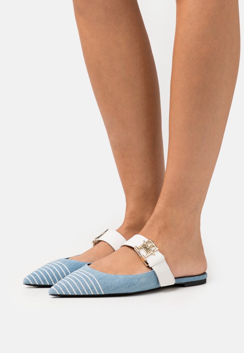 Jambes portant des ballerines bleu pointues à enfiler avec des orteils rayés blancs et des brides blanches ornées de boucles dorées, sur fond blanc.