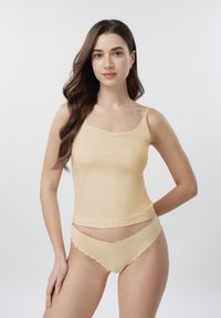 Jeune femme aux cheveux longs bruns portant un caraco beige et une lingerie assortie à bord festonné, debout devant un fond clair uni.