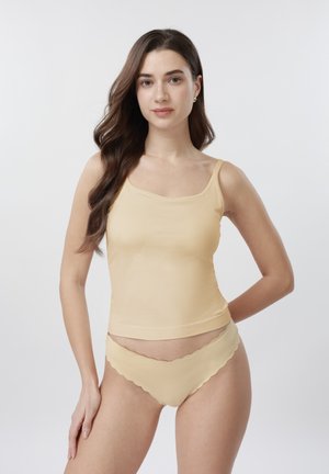 Jauna sieviete ar gariem brūniem matiem, tērpusies bēša krāsas topā un atbilstošā apakšveļā ar vilnainu malu, stāv pret vienkrāsainu gaišu fonu.