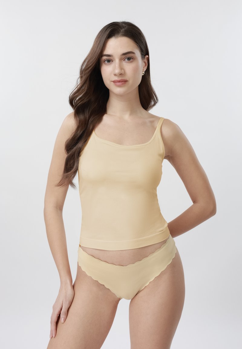 Jeune femme aux cheveux longs bruns portant un caraco beige et une lingerie assortie à bord festonné, debout devant un fond clair uni.