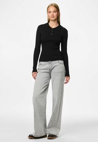 Žena u prilagođenoj crnoj rebrastoj majici dugih rukava s kragnom, svijetlosivim širokim trapericama i crnim cipelama, stoji naslonjena na bijelu pozadinu.