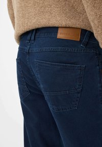 Pantalons en denim bleu marine avec une texture lisse, dotés de deux poches arrière et d'un patch en cuir à la taille portant la mention "EUREX par BRAX."