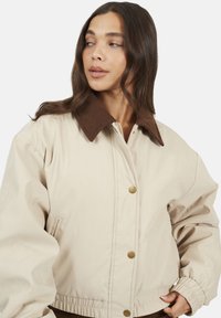 Veste beige en tissu lisse, dotée d'un col en velours côtelé marron, de boutons-pression, de poches latérales et de poignets élastiques pour un ajustement sécurisé.