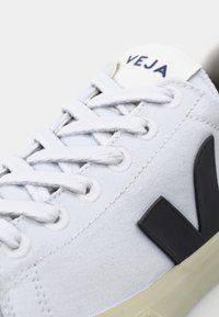 Biele plátěné tenisky s okrúhlou špičkou, šedou gumovou podrážkou a výrazným čiernym logom "V" po boku. Šnúrky a branding v tmavom námorníckom odtieni.