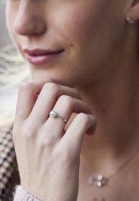 Silaring med en rund, glittrande klar ädelsten monterad på en smal ring, bärs på en hand med ljus hud mot en mjukt suddig bakgrund.