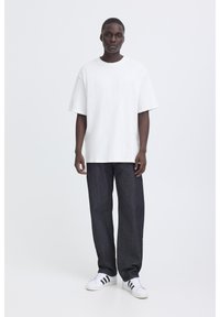 Ung man som står, klädd i en oversized vit T-shirt, mörk löst sittande jeans och vita sneakers med svarta ränder, mot en enkel vit bakgrund.
