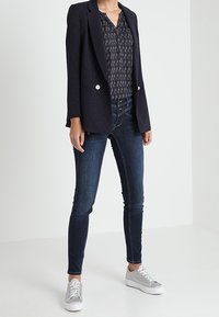 Marinblå blazer med rutmönster över en mönstrad blus, tillsammans med mörka smala jeans och silver sneakers. Blazern har guldknappar.