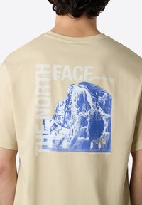 Beige tričko s modrou grafikou hor a textem "THE NORTH FACE", v uvolněném střihu a kulatém výstřihu. Pod tím je nápis "NEVER STOP EXPLORING".