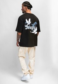 Schwarzes Baumwoll-T-Shirt mit Grafik von zwei weißen Tauben und dem Wort "Dropsi" in stilisierter Schrift auf dem Rücken; kombiniert mit beigen Cargo-Hosen und weißen Turnschuhen.