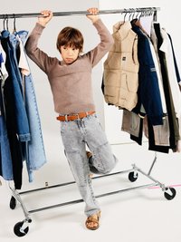 Child in a brown knitted sweater, gray patchwork jeans, and brown sandals, holding onto a clothing rack with various garments hanging.