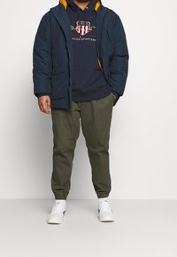 Giacca puffer blu navy con fodera arancione sopra un hoodie blu navy con grafica, abbinata a pantaloni jogger verde oliva e sneakers bianche.